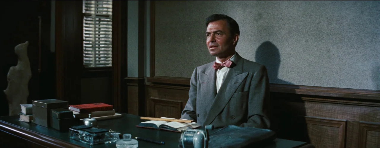 Bigger Than Life (1956) movie still: medium — A man sits behind a wood desk in an office with paneled walls. He wears a suit and bowtie while…