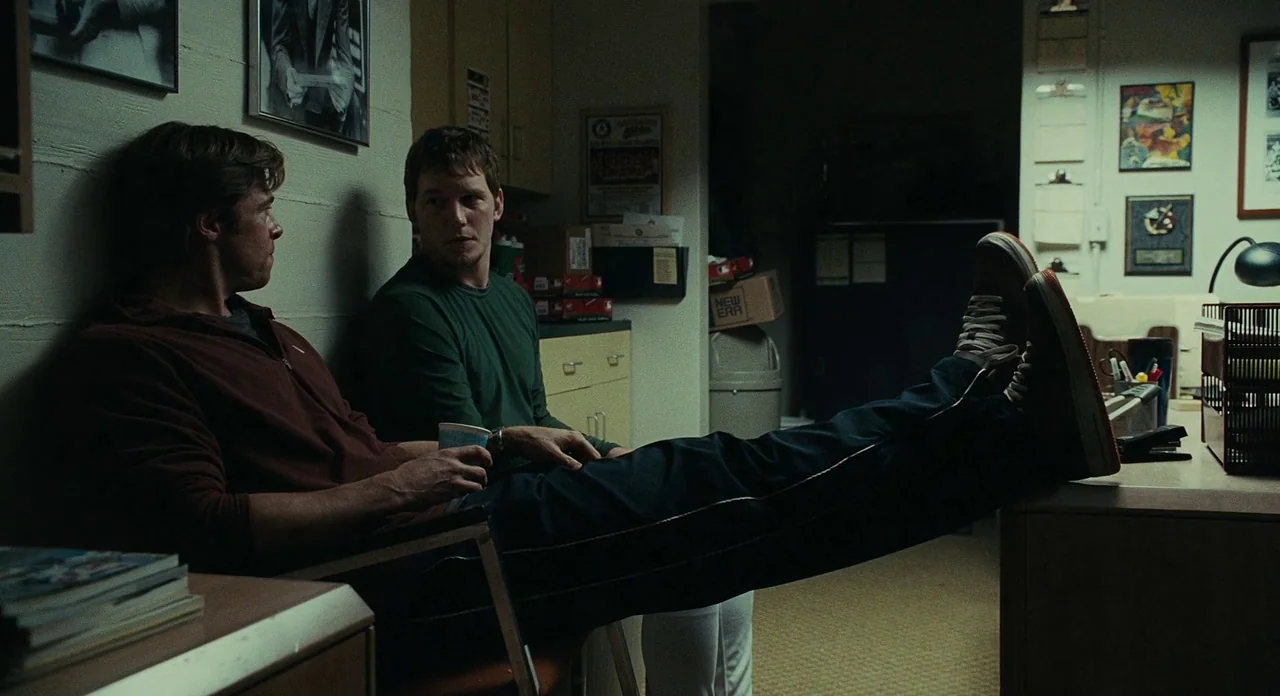 Moneyball (2011) movie still: medium — A utilitarian professional locker room with beige walls and framed athletic posters. Two men sit in chairs…