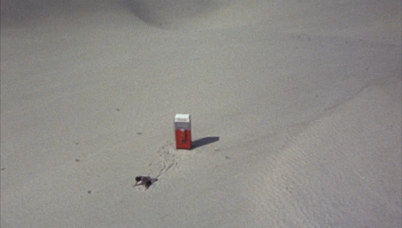 Head (1968) movie still: wide — A vast, empty sandy landscape with rolling dunes under bright, direct sunlight. A lone man crawls on his hands and…