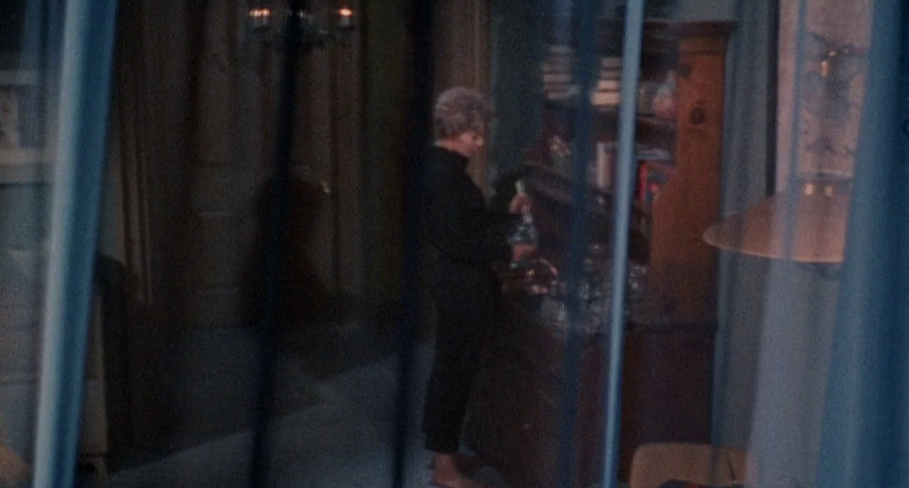 Bell Book and Candle (1958) movie still: medium — A dimly lit room contains a wooden hutch filled with glass items. A woman in dark clothing pours a…