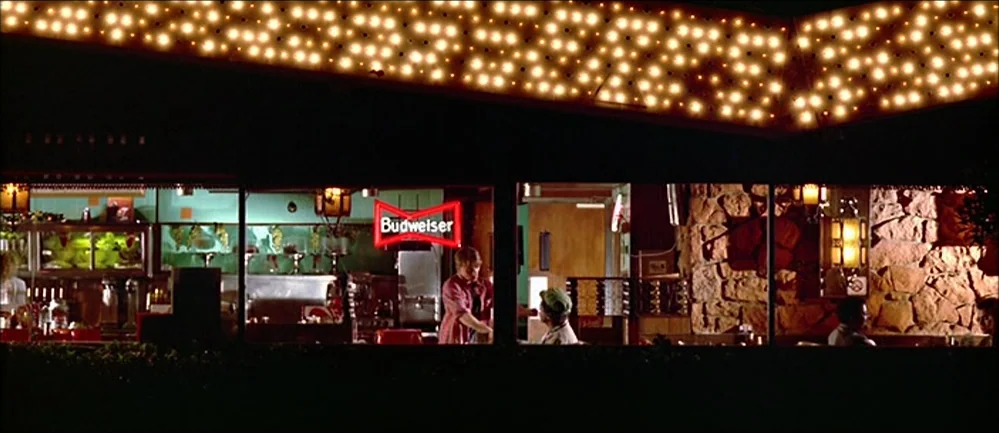 Reservoir Dogs (1992) movie still: wide — A long, single-story building at night featuring a row of glowing exterior lights. People are visible…