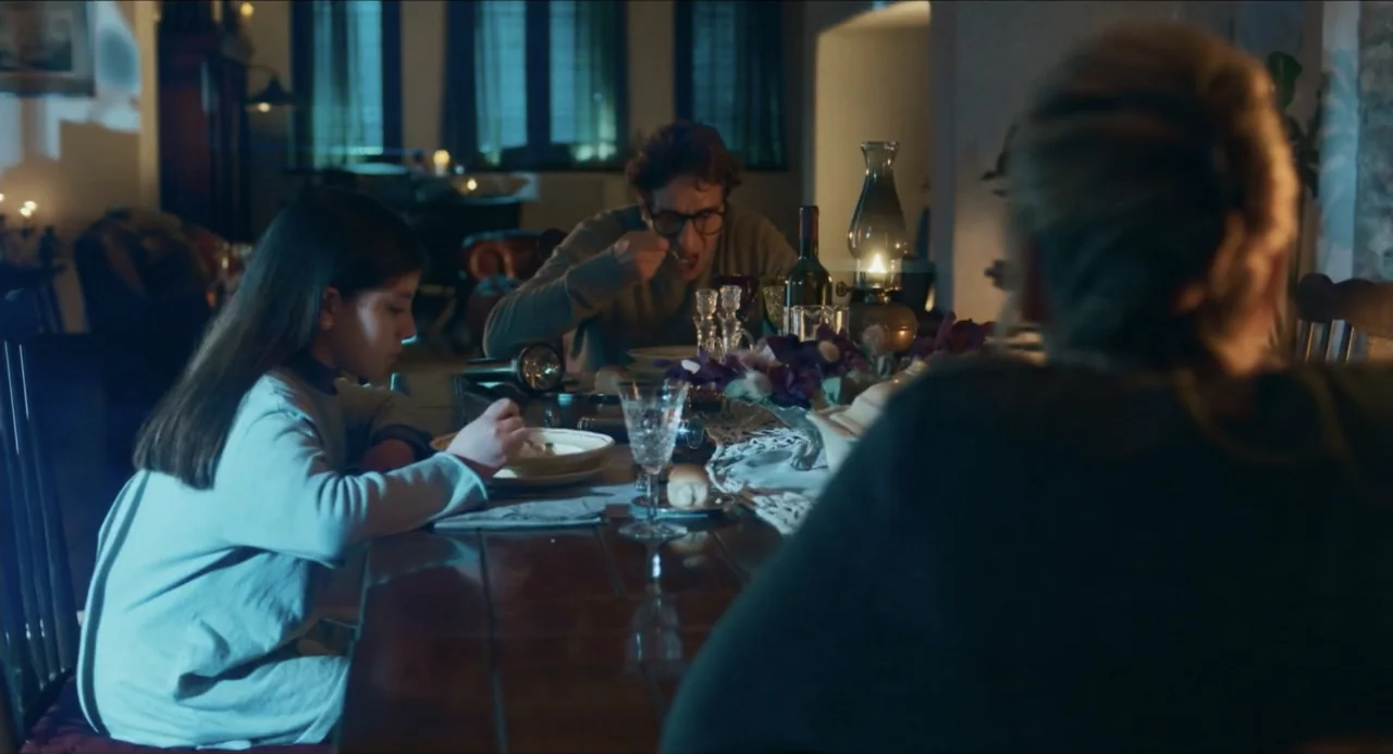 Echoes Of A Crime (2025) movie still: medium — A dark dining room features a long wooden table set for an evening meal. A child and a man sit…