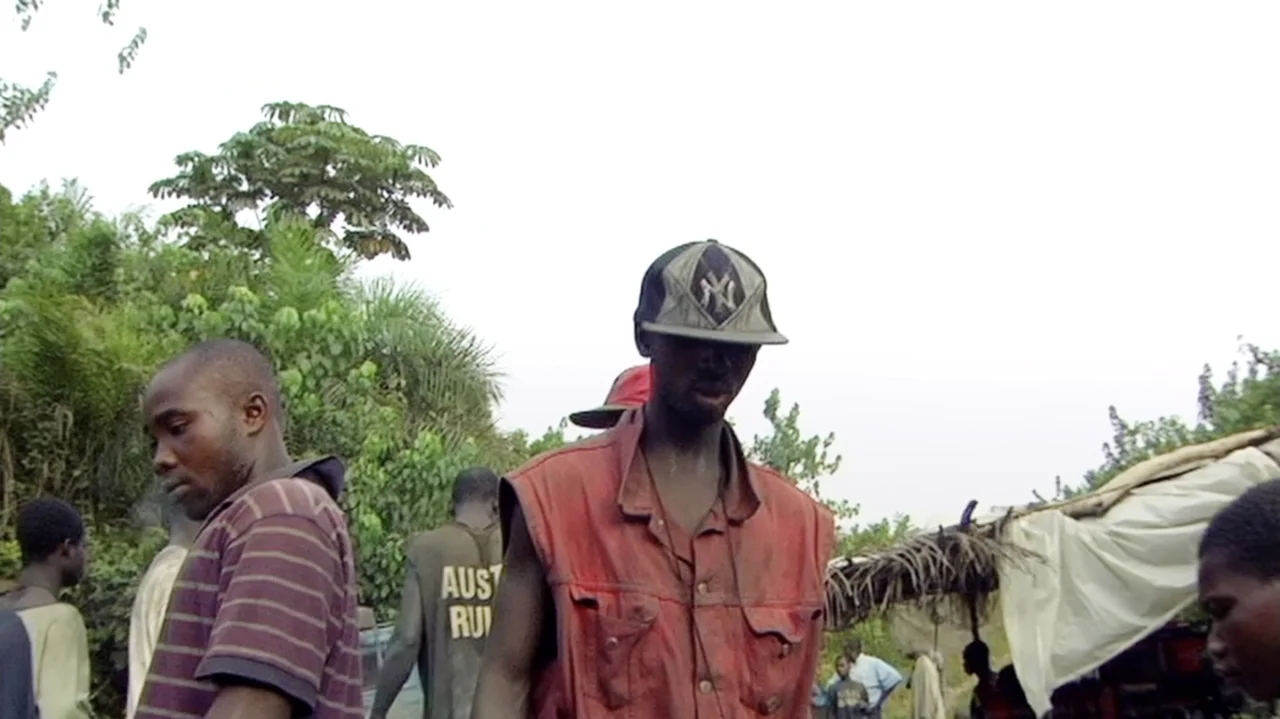 The Money Stone (2018) movie still: medium — A group of people stands on a dirt path surrounded by dense tropical foliage. A man wearing a red vest…