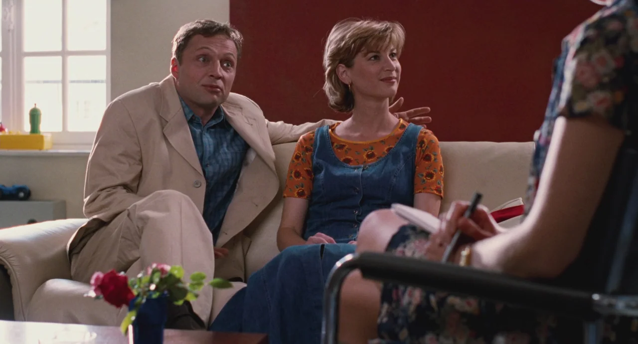 Ma Vie En Rose (1997) movie still: medium — A man and woman sit side-by-side on a cream-colored couch in an office. They are turned toward an unseen…