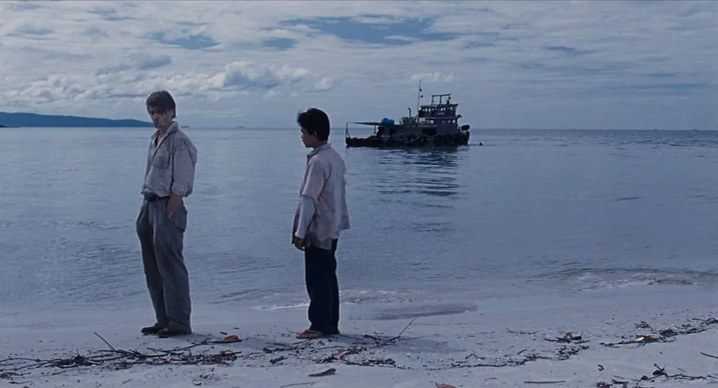 Almayer’s Folly (2011) movie still: fullbody — A quiet coastal landscape features a sandy beach under a thick overcast sky. Two men stand near the…