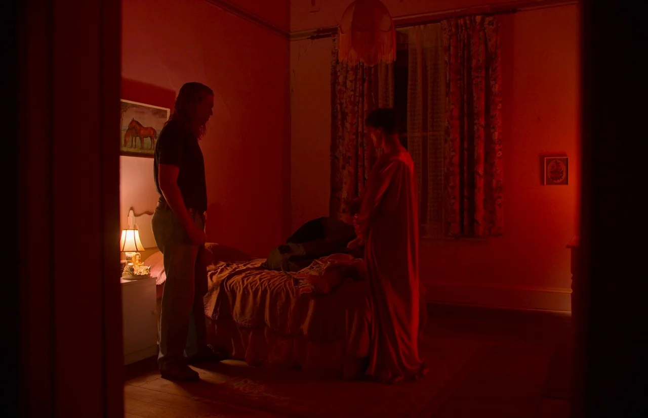 Nitram (2021) movie still: medium — A dimly lit room is illuminated by deep red light and contains a bed. A long-haired man stands by the bed facing…