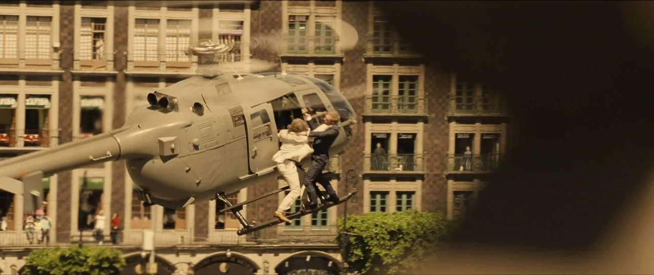 Spectre (2015) movie still: wide — A grey helicopter flies low above a city square lined with multi-story historic buildings. Two men in formal…