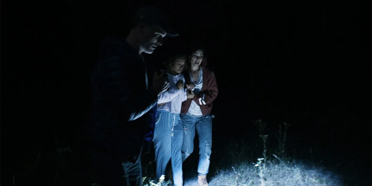 Demonic (2015) movie still: medium — Three individuals move across a dark, grassy field at night under artificial illumination. A man in a cap leads…