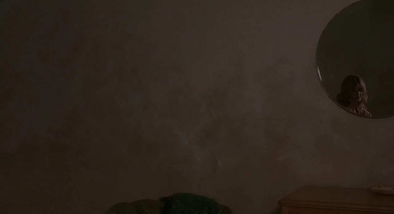 Certain Women (2016) movie still: wide — A dimly lit room features a large, dark, textured wall and a circular mirror. A woman's face is visible…