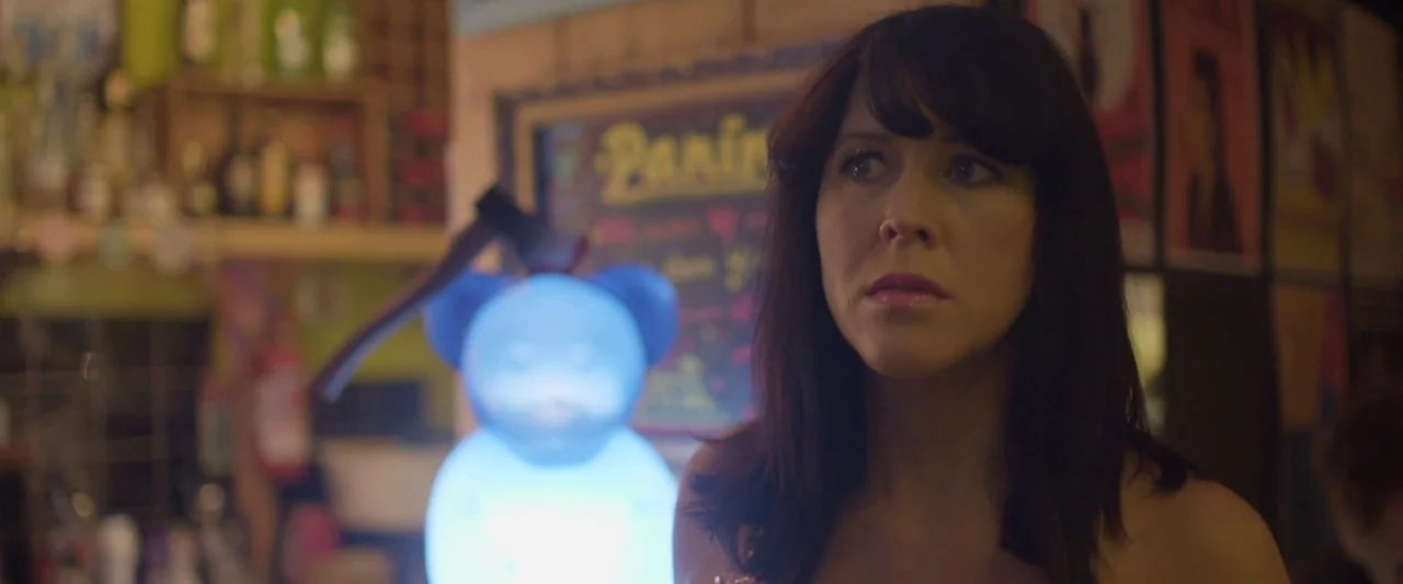 Prevenge (2016) movie still: medium — An interior space with a dimly lit shelf of bottles and posters on walls. A woman stands in the foreground…