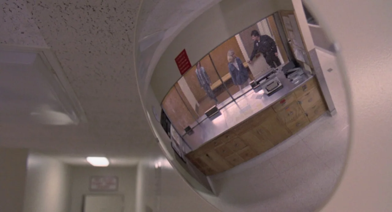 River’s Edge (1986) movie still: wide — A hospital hallway features a convex security mirror reflecting a nearby reception desk area. Three people…