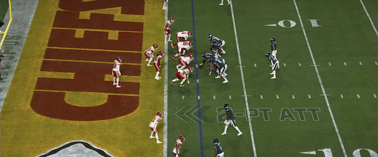 Kelce (2023) movie still: wide — A high-angle view of a professional football field marked with white yard lines and team logos. Players in red and…