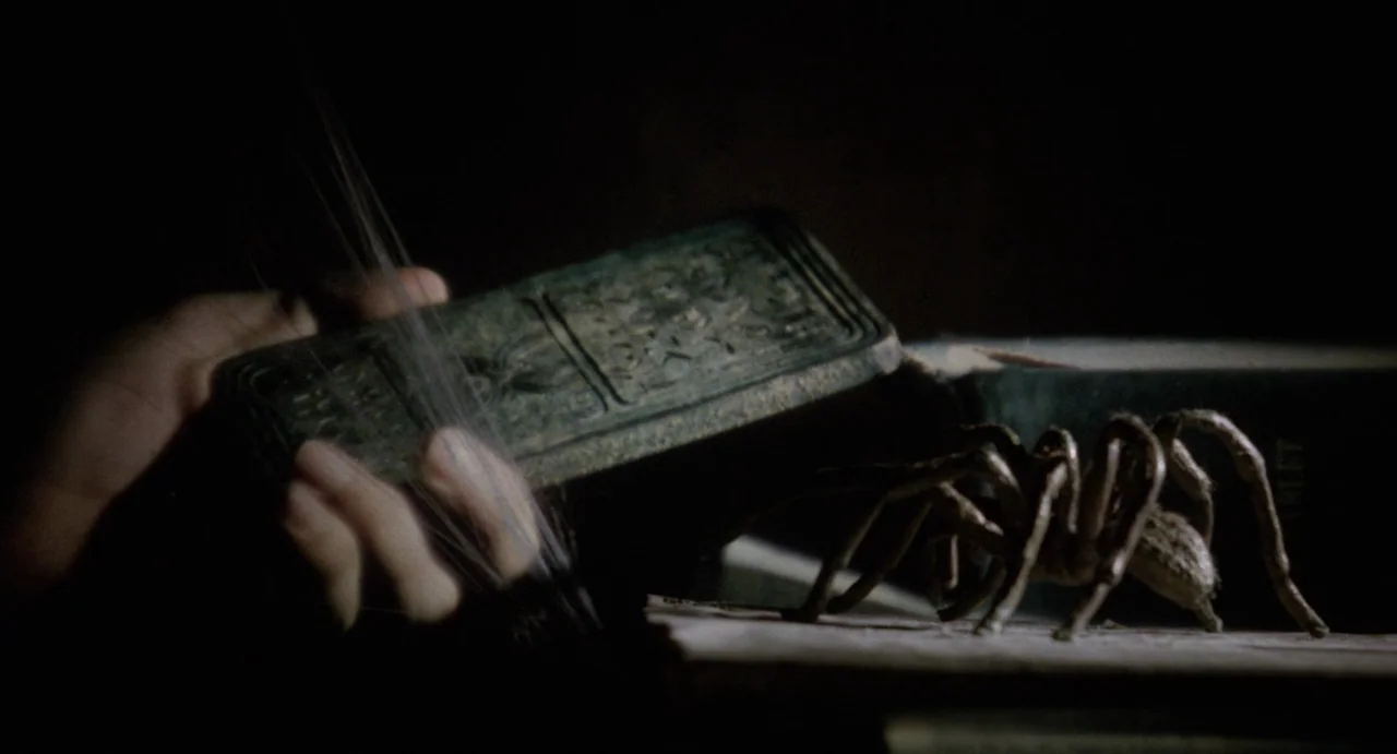 The Spider Labyrinth (1988) movie still: closeup — A spider traverses the page of an open book in a dark room. A hand holds a small embossed…