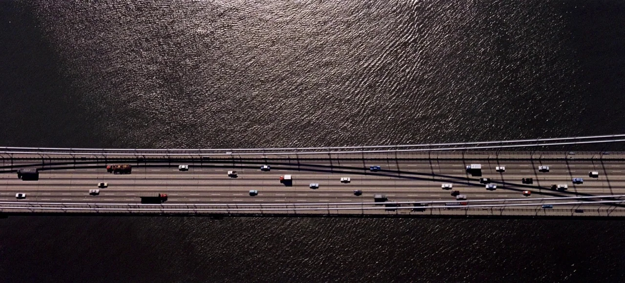 West Side Story (1961) movie still: establishing — A high-angle view shows a long highway bridge spanning across dark reflective water. Numerous…