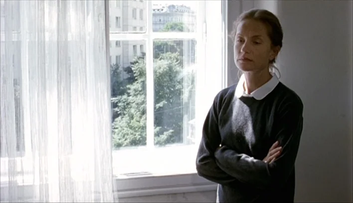 The Piano Teacher (2001) movie still: medium — A spare interior space with white walls and a large, multi-paned glass window. A woman stands in…