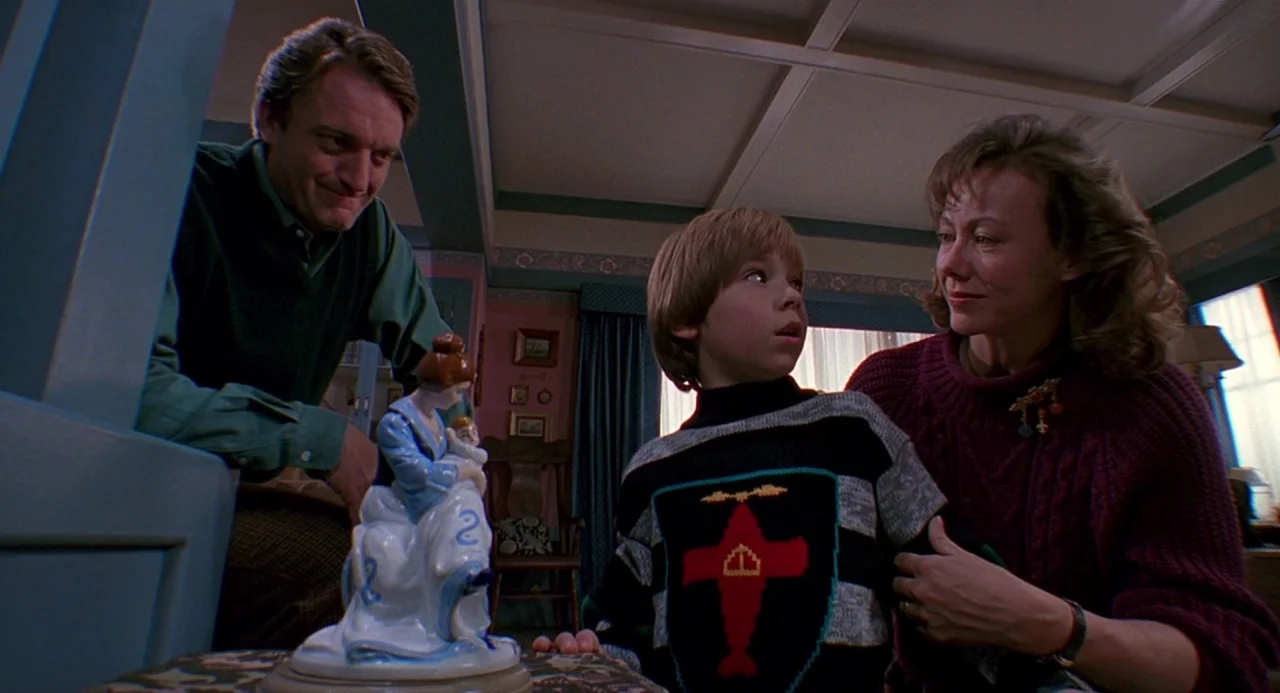 Child’s Play 2 (1990) movie still: medium — A family sits together in a 1990s domestic space with floral wallpaper and moldings. The man, woman, and…
