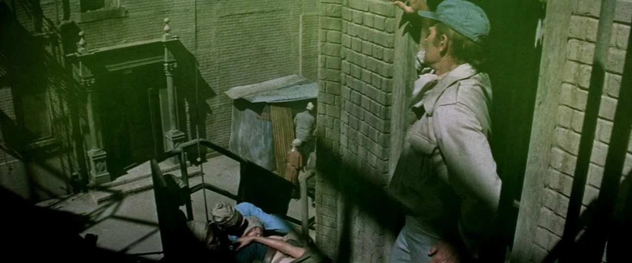 Soylent Green (1973) movie still: wide — An urban alleyway features brick walls, metal fire escapes, and a small makeshift shelter. A man stands on…