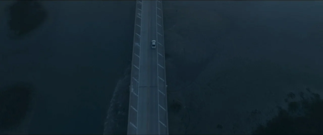 Offseason (2022) movie still: establishing — A long bridge extends over dark, calm water at night. A lone white sedan drives down the center of the…