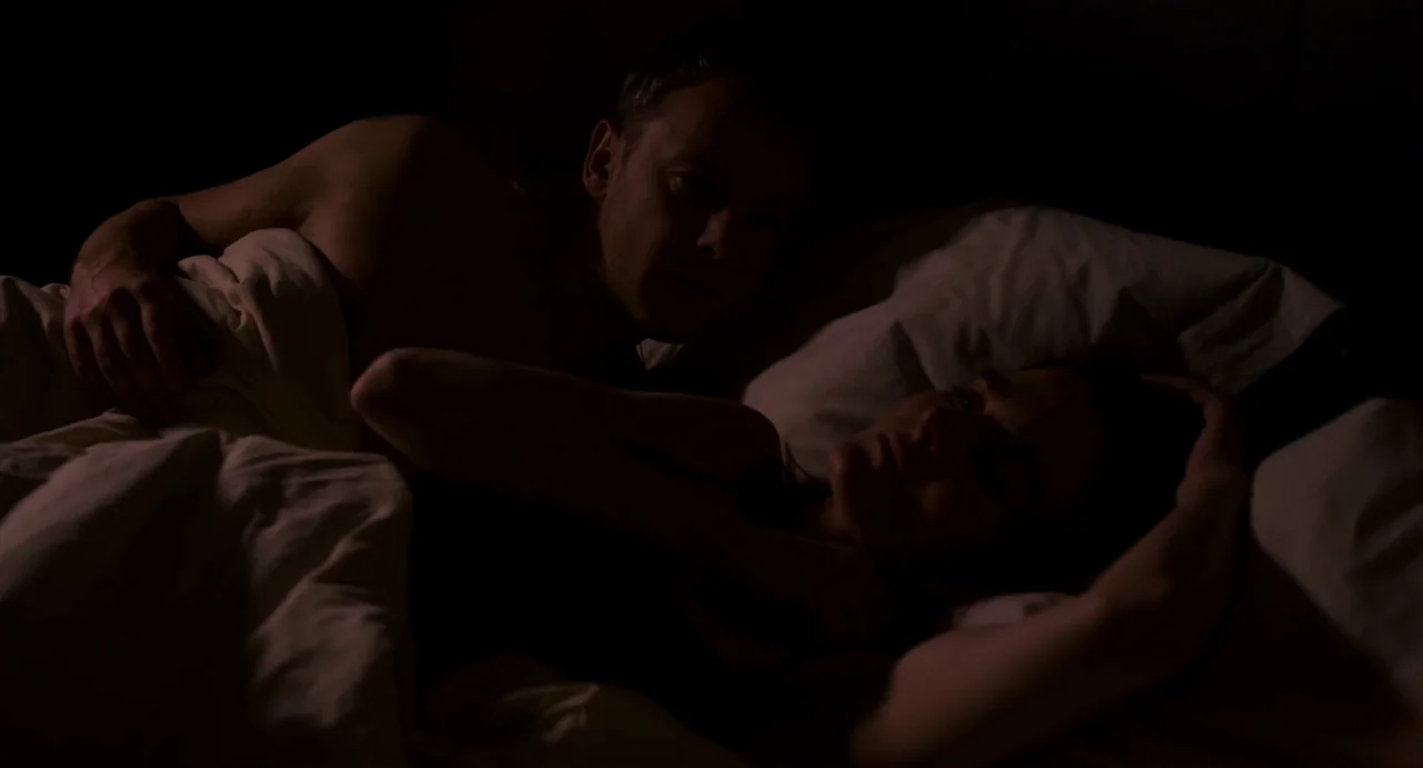 Everyday (2012) movie still: medium — A dark, dimly lit space contains two people lying close together in a bed. They are resting under white…