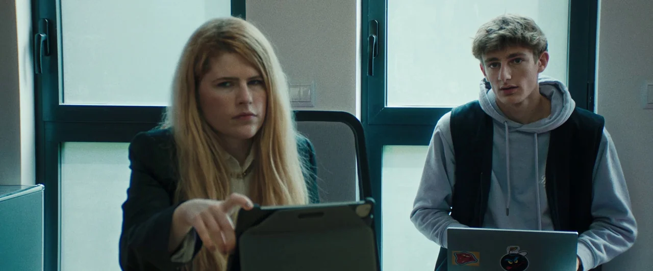 Vincent Must Die (2023) movie still: medium — A modern workspace or classroom with large glass windows showing bright daylight outside. A woman in a…