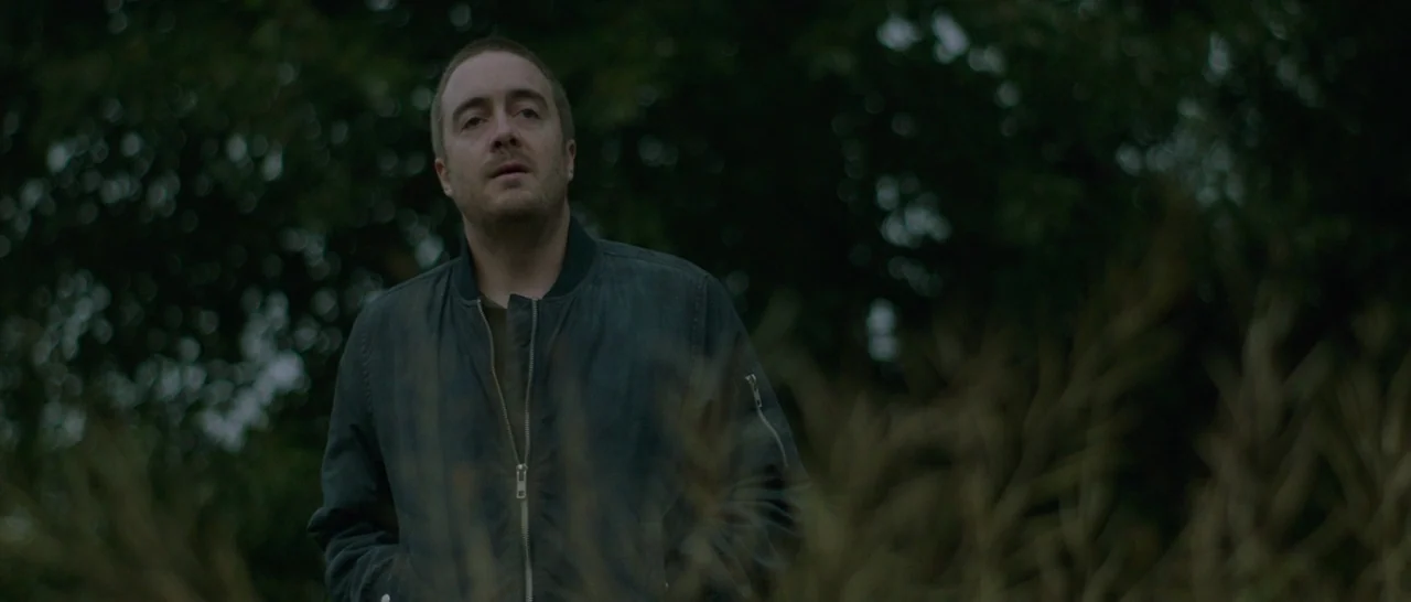 Green Room (2015) movie still: medium — A man stands in an outdoor area covered in tall, dry, wild grasses. Dense green trees are visible in the…