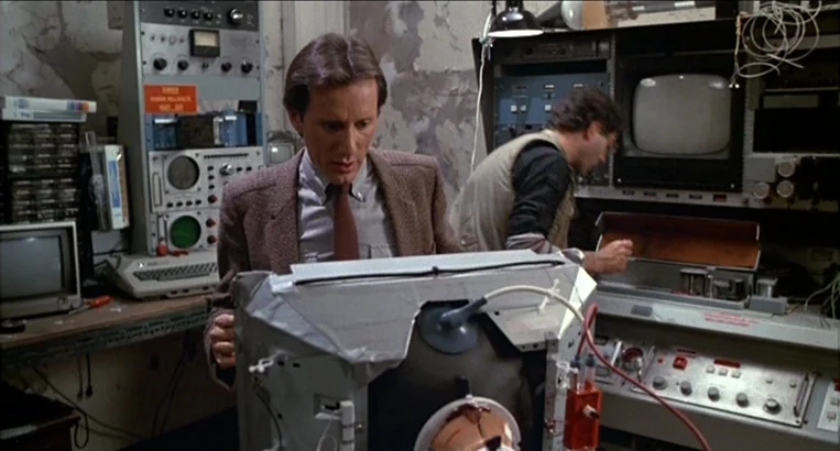 Videodrome (1983) movie still: medium — A cluttered technical laboratory featuring peeling wallpaper and racks of vintage 1980s electronic…
