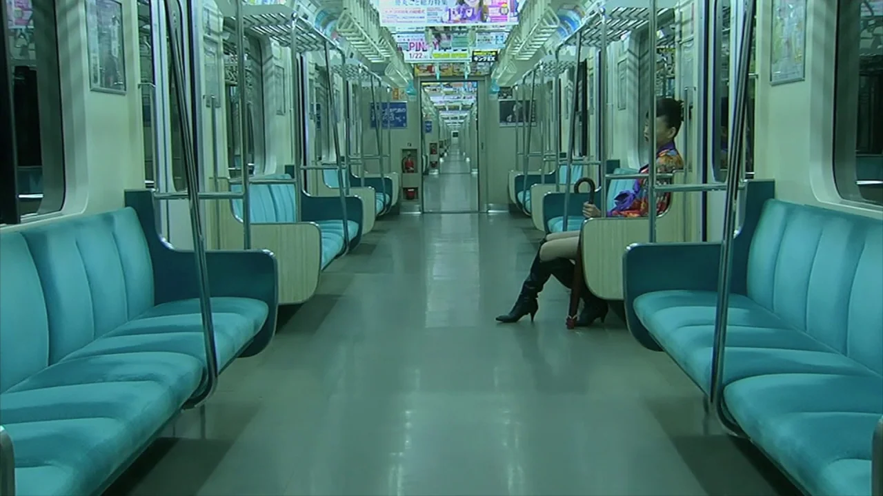 Tokyo Gore Police (2008) movie still: wide — A nearly empty train car interior features two rows of blue-green cushioned bench seating. A lone woman…