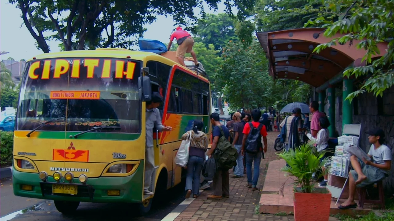 Merantau (2009) movie still: wide — A yellow and green passenger bus is parked by a roadside concrete shelter. Commuters prepare to board the…