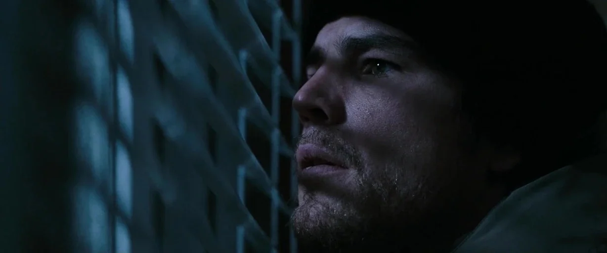 30 Days of Night (2007) movie still: closeup — A dimly lit room interior features a person peering through slatted window blinds at night. The man…