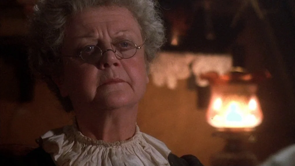 The Company of Wolves (1984) movie still: closeup — An interior space is dimly lit by a glowing amber oil lamp in the background. An elderly woman…