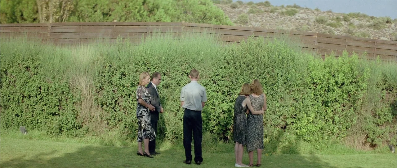 Dogtooth movie still: wide — A group of five people stands on a lawn before a tall green hedge. A wooden fence runs horizontally across the top of…