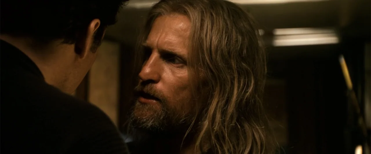 2012 (2009) movie still: closeup — A dimly lit indoor space with shadows obscuring the surrounding environment details. A bearded man with long hair…