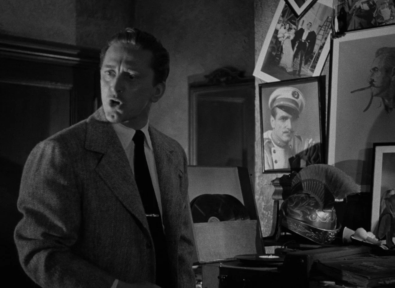 The Bad and the Beautiful (1952) movie still: medium — A dimly lit study decorated with framed photographs and a decorative historical helmet. A man…