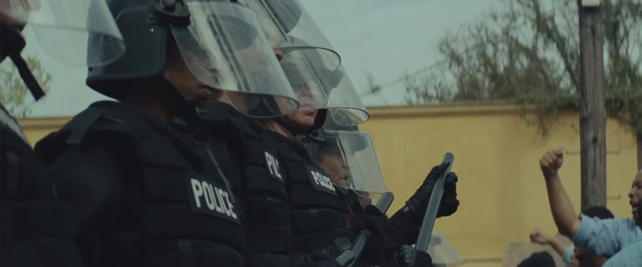 Queen & Slim (2019) movie still: medium — A line of police in black riot gear stands against an exterior yellow wall. Several officers wear…
