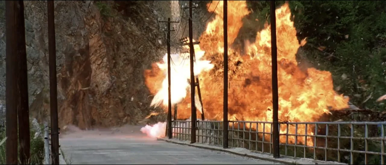 The Peacemaker (1997) movie still: wide — An asphalt road runs alongside a steep rock face and a metal railing. A massive explosion of fire and…