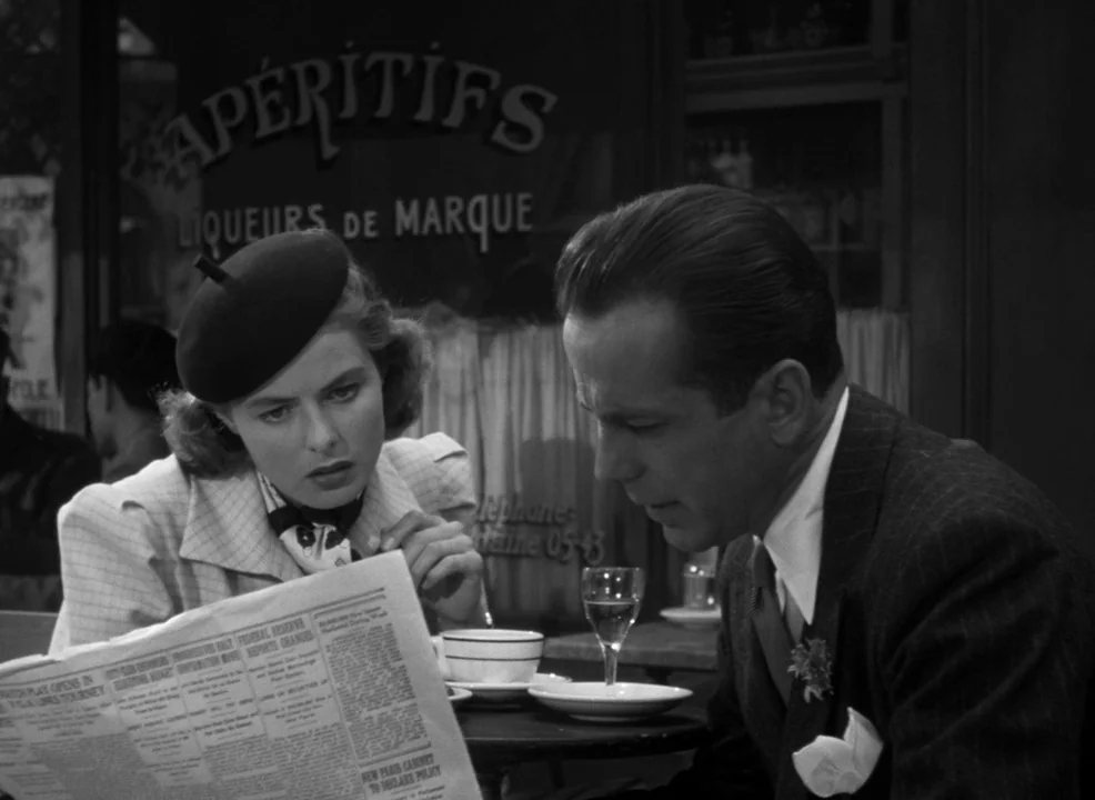 Casablanca (1942) movie still: medium — A sidewalk cafe terrace features a window behind the subjects with painted French text. A woman in a beret…