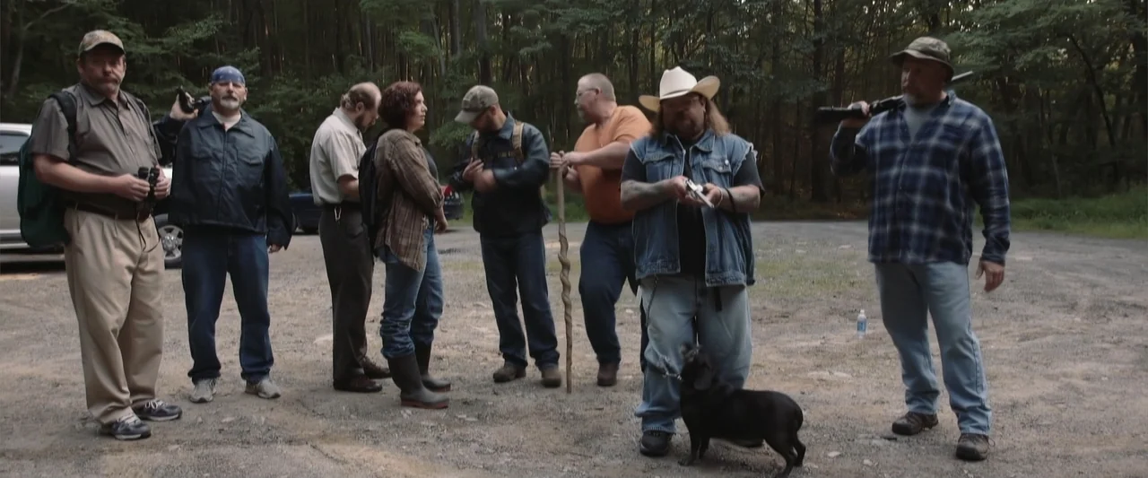 Darkness Waits (2020) movie still: fullbody — A gravel clearing is bordered by dense green woods under natural daylight. A group of several adults…