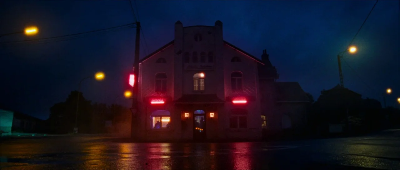 Inexorable (2021) movie still: wide — A two-story building sits at a street corner on a wet night. Vibrant red neon signs glow against the dark…
