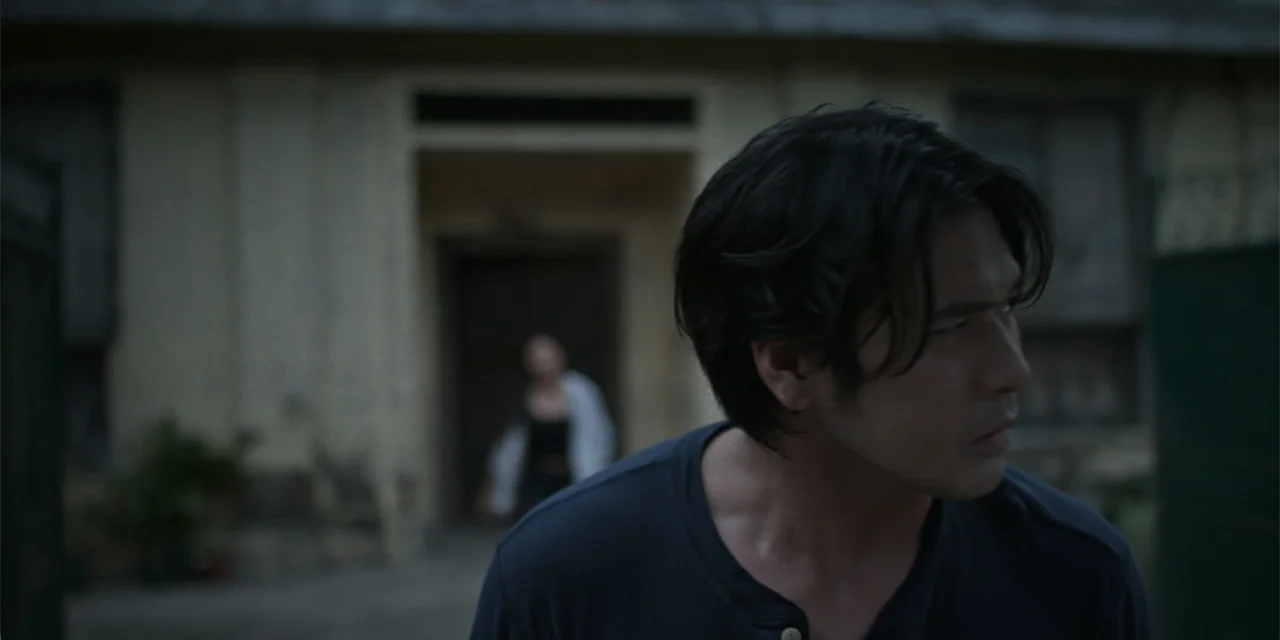 Nokturno (2024) movie still: medium — A man stands in the foreground in front of a blurred building entrance. A woman is visible out of focus in the…