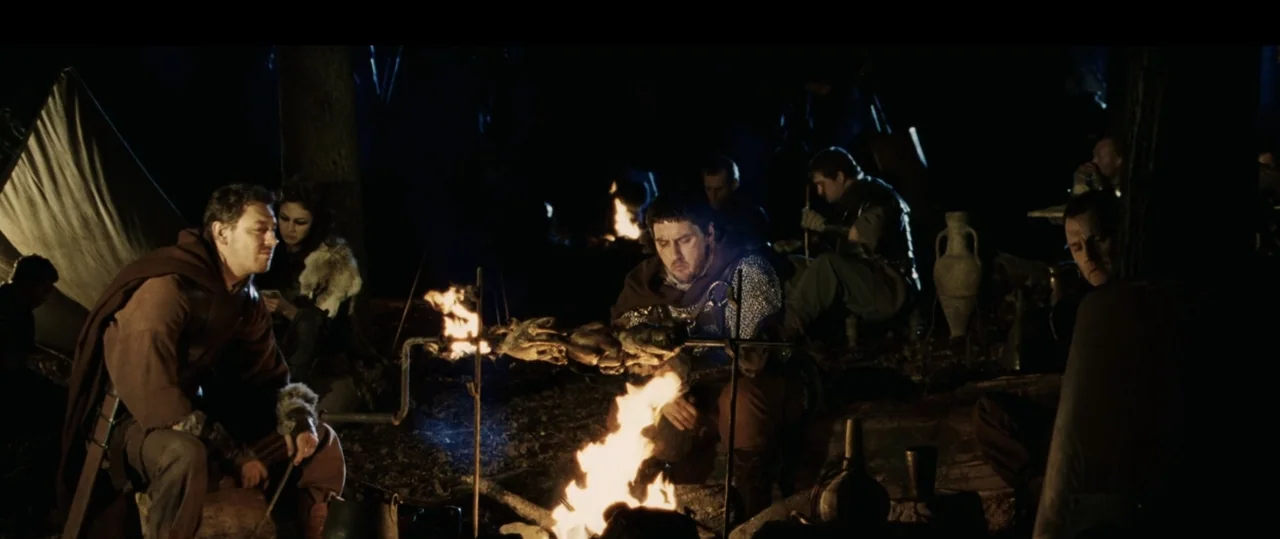 Centurion (2010) movie still: wide — A group of travelers rests around a crackling fire in a dark wooded encampment. A man in chainmail rotates meat…