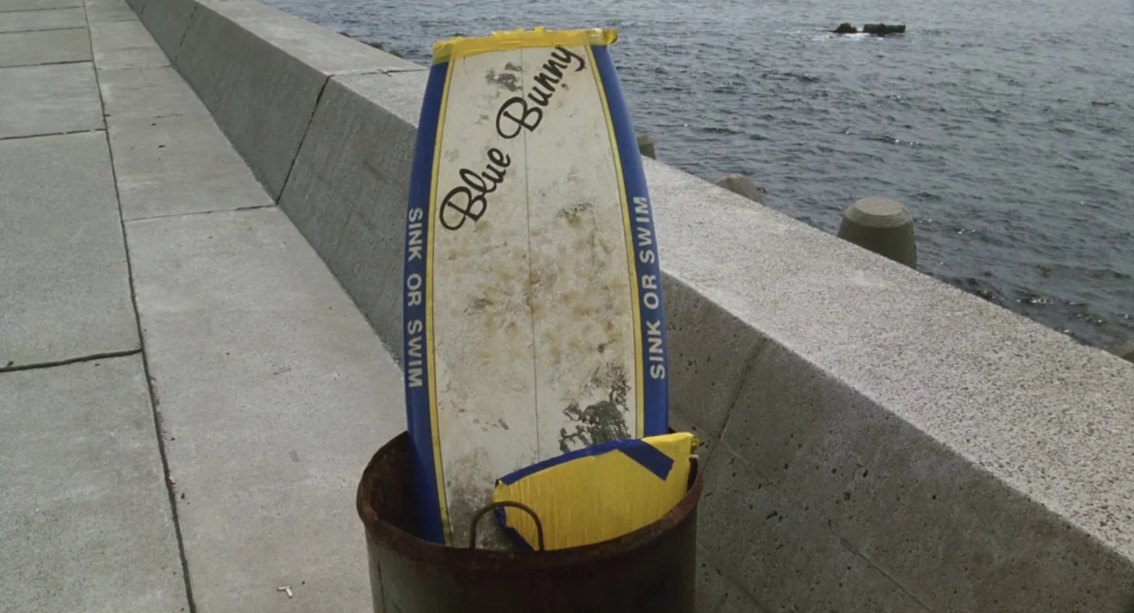 A Scene at the Sea (1991) movie still: medium — A worn surfboard with text stands upright inside a rusted barrel near the ocean. The backdrop…
