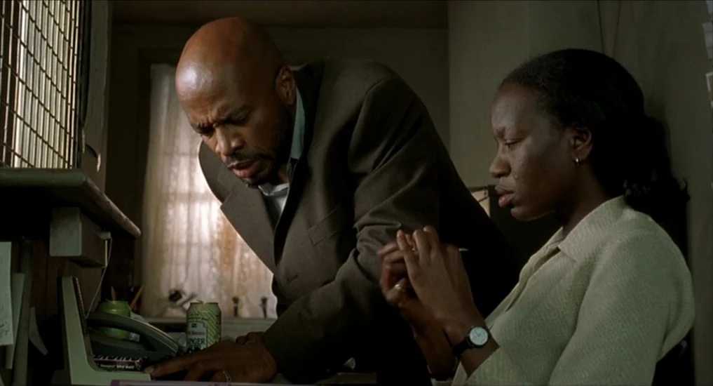 All or Nothing (2002) movie still: medium — A cramped, dimly lit workspace features a wooden desk with papers and an telephone. A bald man leans…