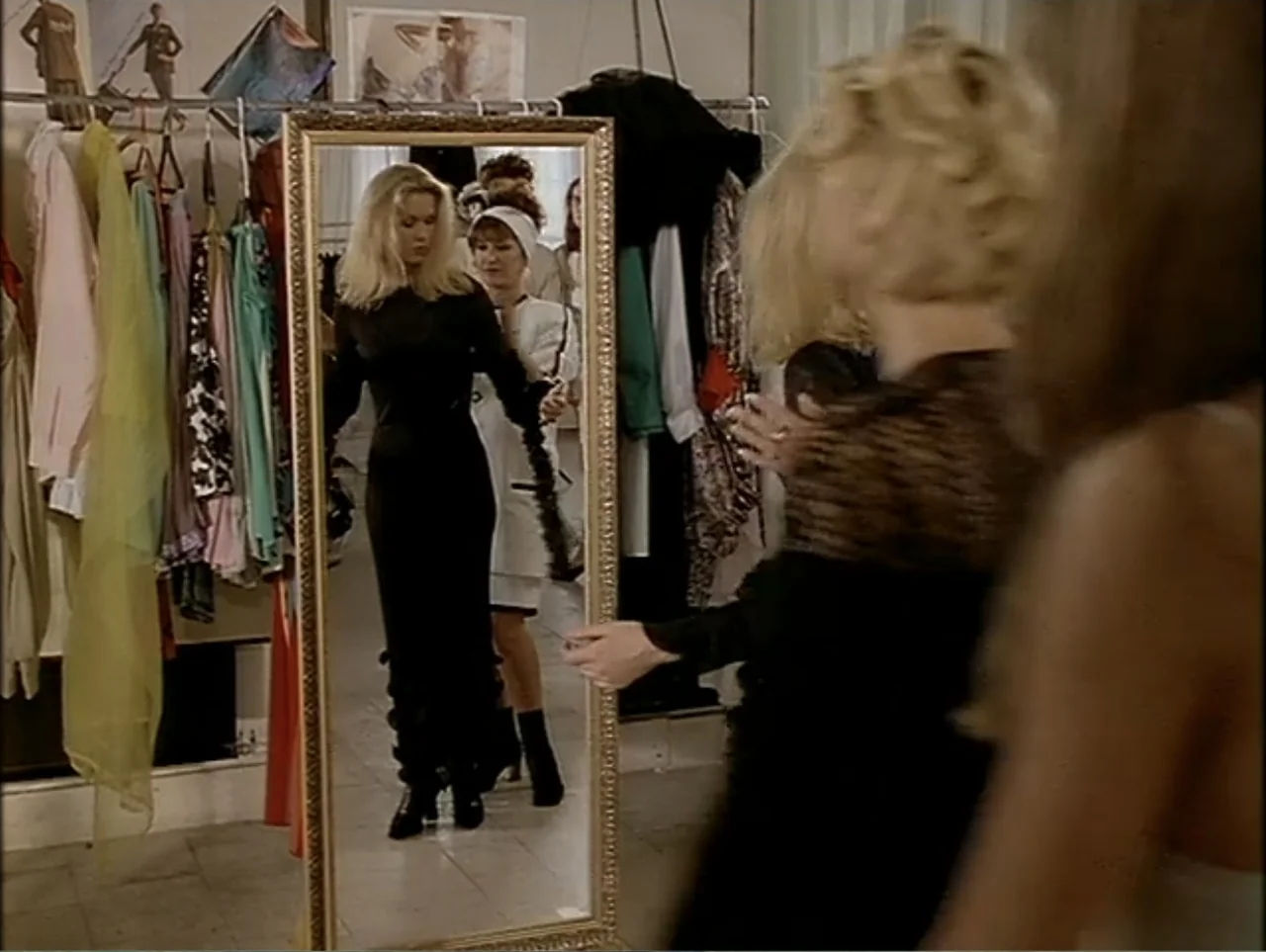 A Change of Place (1994) movie still: medium — A dressing room contains a large rectangular mirror and racks filled with diverse garments. A woman…