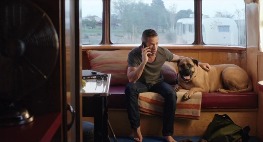 Results (2015) movie still: medium — A small camper interior features wooden walls, built-in benches, and large rear windows. A man sitting on a…