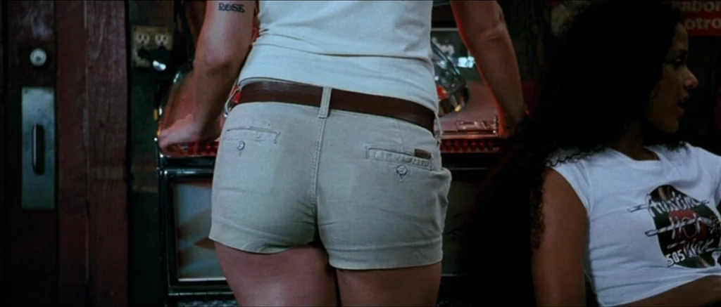 Death Proof movie still: medium — A dimly lit room features a jukebox against a wooden wall. Two women stand or sit near the machine in a casual,…