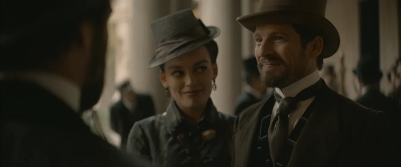 Eiffel (2021) movie still: medium — A stone colonnade serves as the background for a gathering of people in period attire. A man and a woman stand…