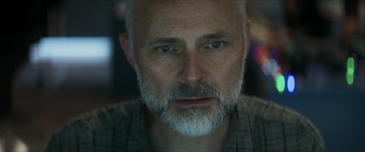 Last Breath (2025) movie still: closeup — A dark indoor space with blurred colorful lights visible in the background. A middle-aged man with grey…