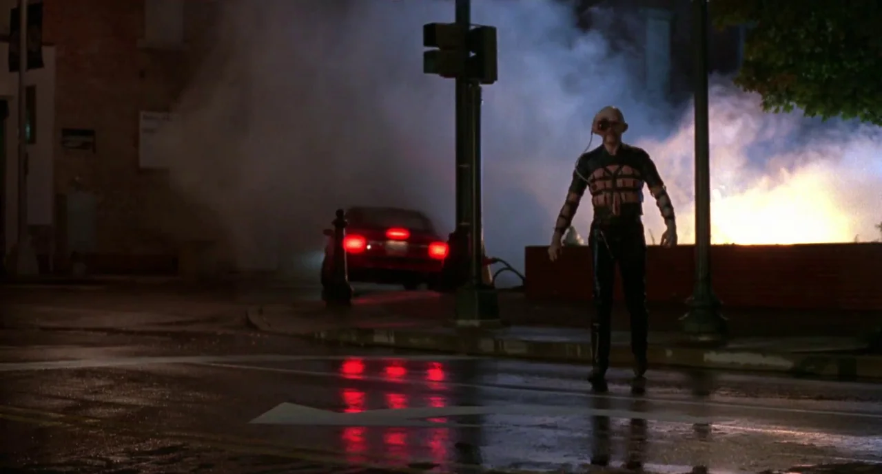 Hellraiser III: Hell on Earth movie still: fullbody — A wet asphalt intersection at night features a man standing in the foreground. A red car is…