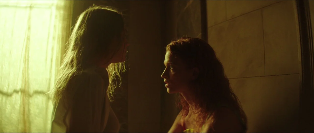 Braid (2018) movie still: medium — A dimly lit interior with a bright window source illuminating two women facing each other. One woman appears to…