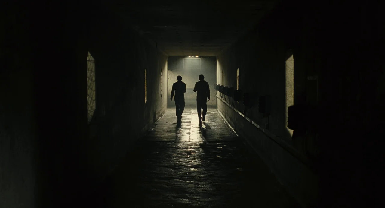 The Double (2013) movie still: wide — A long concrete corridor is illuminated by a single bright light at the end. Two men in dark suits walk away…